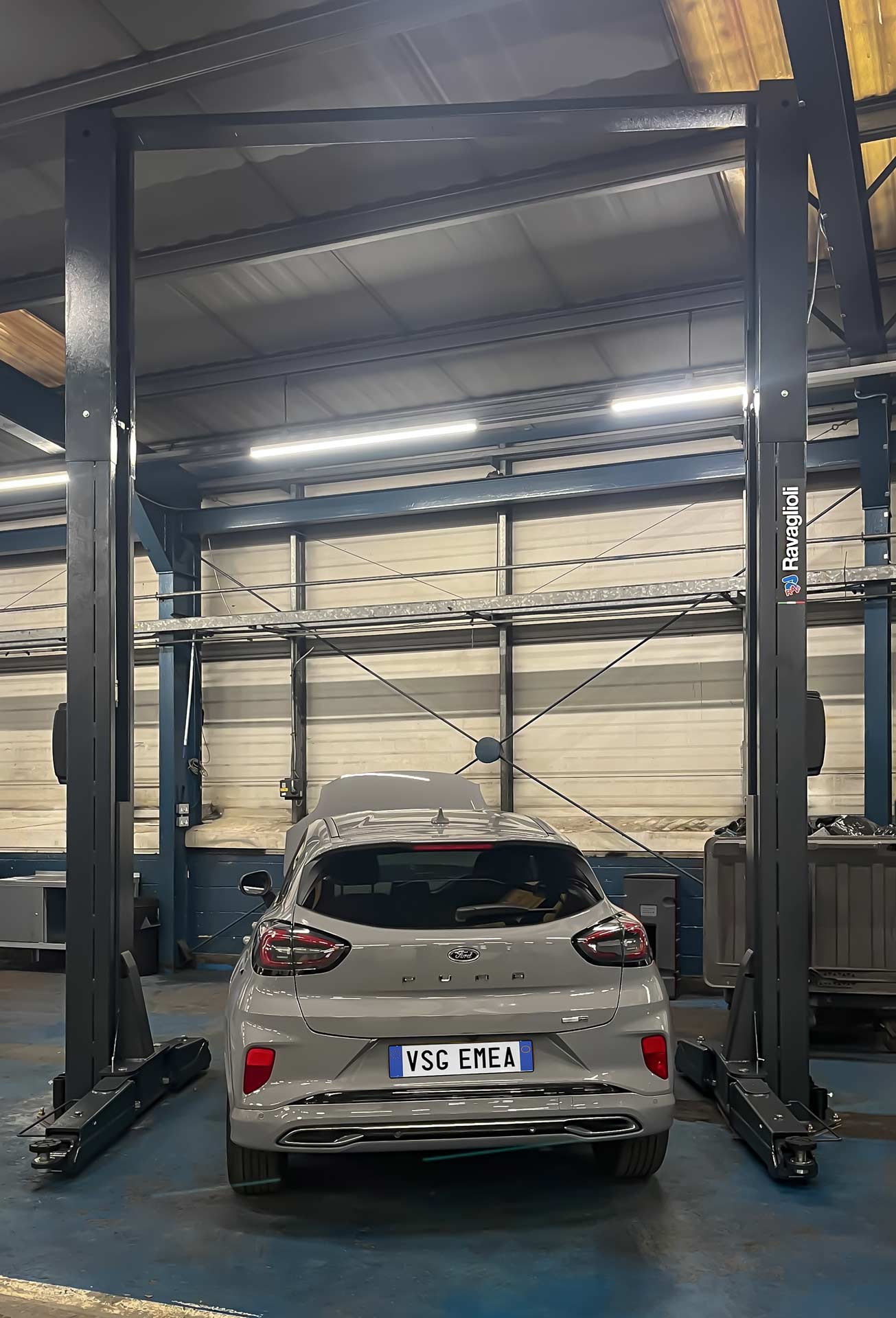 Gray Ford Puma is serviced in a workshop parked between the columns of a black Ravaglioli lift