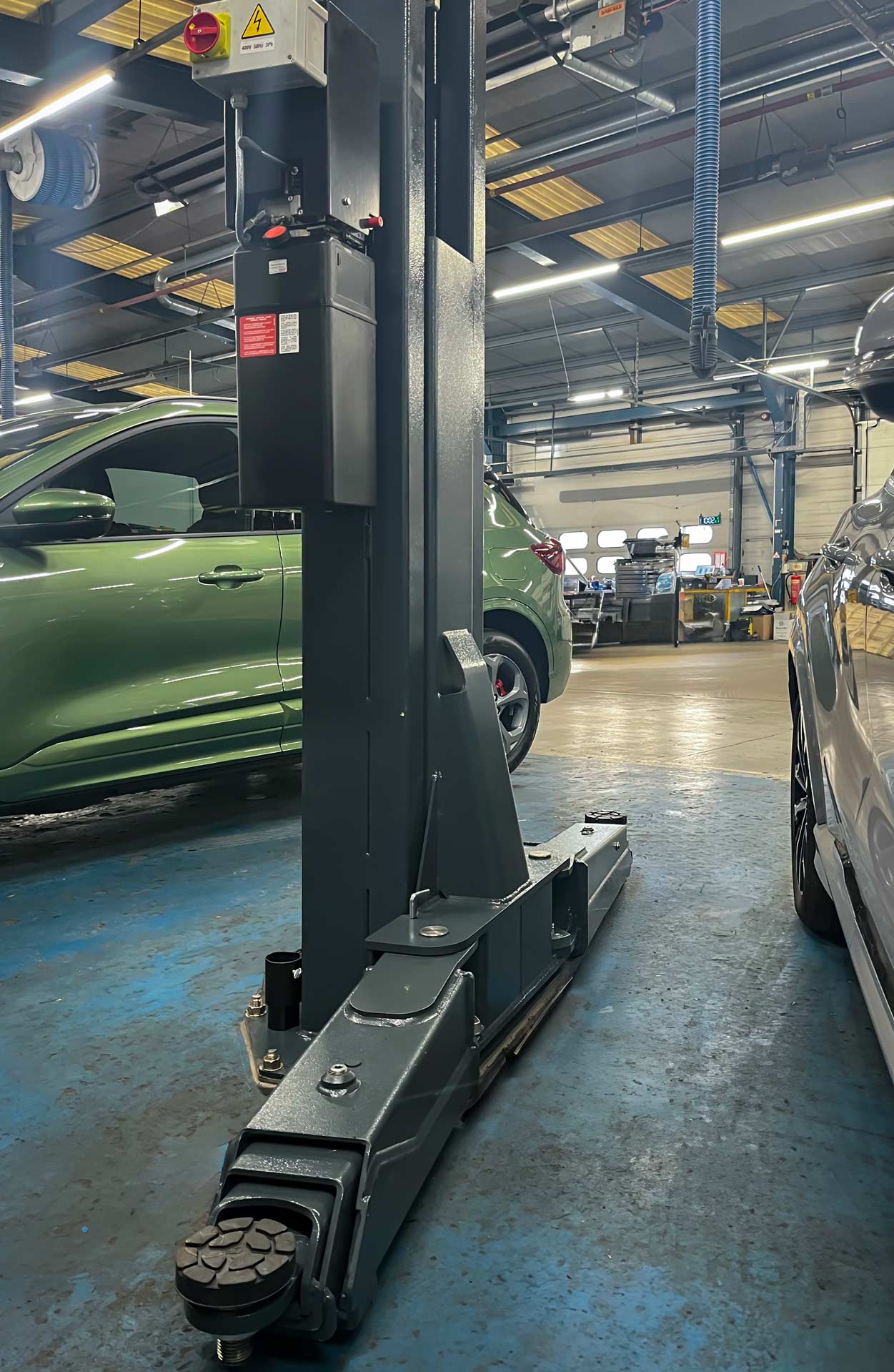 Two arms of a black machine workshop car lift with green car in the background